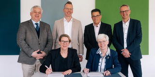 Auf dem Foto sieht man insgesamt sechs Personen in die Kamera lächeln. Zwei von den sechs Personen sitzen an einem Tisch und unterzeichnen ein Dokument.
