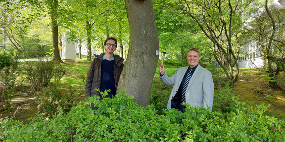 Zwei Männer stehen lachend vor einem Baum, an dem ein Vogelhaus hängt.