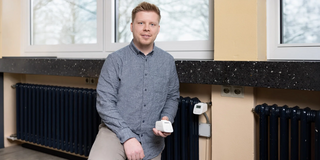 Ein blonder kurzhaariger Mann steht vor einem Heizkörper und hält ein digitales Heizkörperthermostat in der Hand.