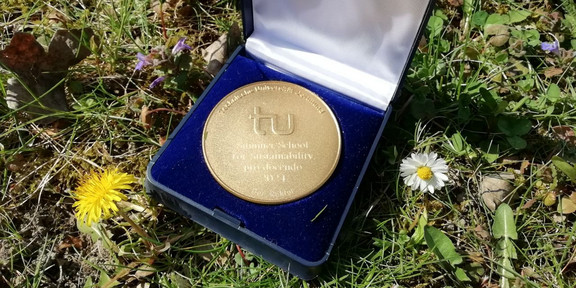 Die Medaille zur Auszeichnung der Summer School for Sustainability mit dem Lehrpreis 2024 der TU Dortmund liegt im Gras umgeben von eineigen Blüten