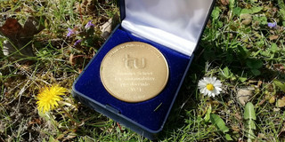 Die Medaille zur Auszeichnung der Summer School for Sustainability mit dem Lehrpreis 2024 der TU Dortmund liegt im Gras umgeben von eineigen Blüten