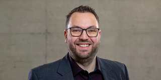 Foto: Ein Mann mit kurzem dunklem Haar steht vor einer grauen Wand und lächelt in die Kamera.