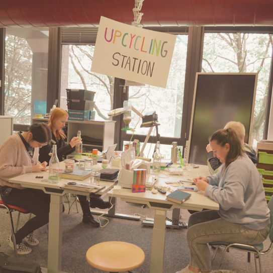 Photo of four people around a double table working on various upcycling projects. A colorful “Upcycling station” sign hangs above the tables.