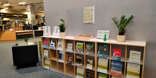 Regal mit Büchern, Pflaanzen und einer Box mit Saatguttüten. Im Hintergrund ein Schalter der Universitätsbibliothek.