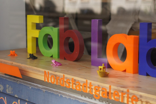 Durch eine Fensterscheibe sieht man 3D gedruckten bunte Buchstaben Fab Lab. Unter Fab Lab steht Nordstadtgalerie auf der Fensterscheibe.