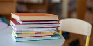Auf dem Bild sieht man einen Bücherstapel auf einem Tisch liegen. Neben dem Tisch steht ein Stuhl.