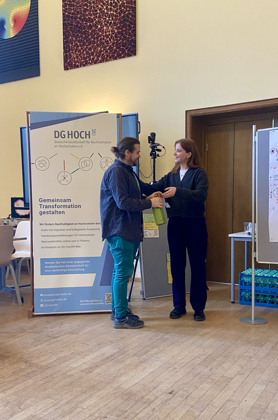 The network manager of INNO4BNE Daria Humburg presents the 1st place prize to the coordinator of studium oecologicum Marvin Maikler for the presentation of the poster on the topic of Summer School for Sustainability as part of a gallery walk.