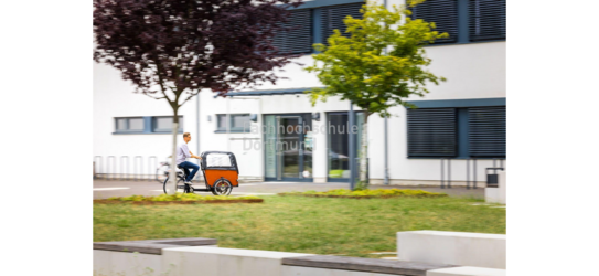 Ein Mann fährt mit einem Lastenrad über den Campus der Fachhochschule Dortmund. Man sieht ein Gebäude und zwei Bäume.