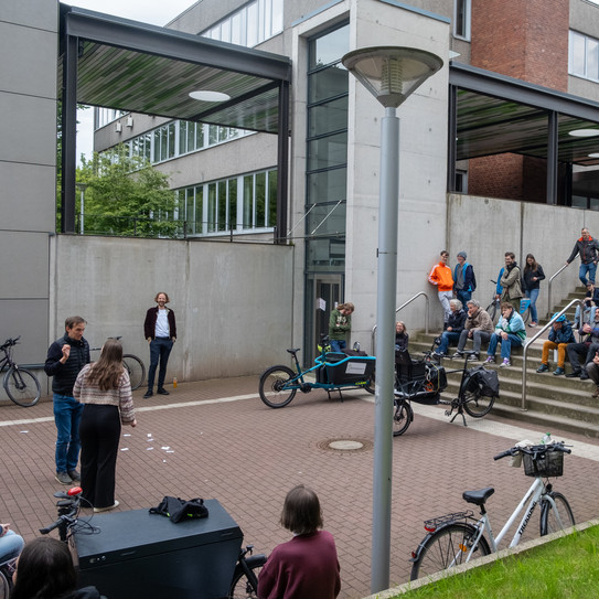 Eine Gruppe von Menschen sitzt auf Treppenstufen und beobachtet eine Veranstaltung im Außenbereich, umgeben von Fahrrädern und moderner Architektur.