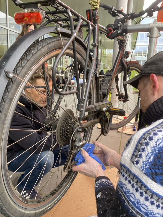 The picture shows a bicycle hanging on a repair rack. On the right side of the bicycle, a man is cleaning the bicycle chain. A man is squatting on the left side of the bicycle.