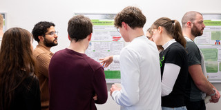 In the center of the picture, four young adults can be seen from the side and from behind, looking together at a poster attached to a white wall. On the left in the foreground, a person with a black top and brown hair can also be seen from behind. To the right of the people, a male person is standing sideways in front of another poster with his back to the other group.