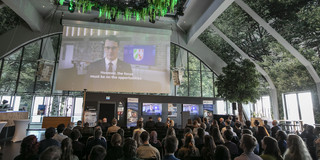 Foto: Ein Veranstaltungssaal im Dortmunder U mit begrünter Decke. Vor dem Publikum wird eine Videobotschaft von Ministerpräsident Wüst abgespielt.