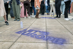 Auf den gepflasterten Boden ist vor einer Gruppe von Beinen im Hintergrund in dunkellila der folgende Text aufgesprüht: „Let's Europe“.