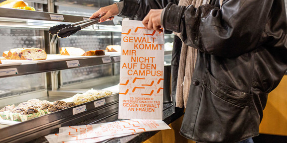 Eine Person nimmt sich aus einer Selbstbedienungstheke ein Stück Kuchen. Sie hält eine Tüte mit der Aufschrift "Gewalt kommt mir nicht in die Tüte".