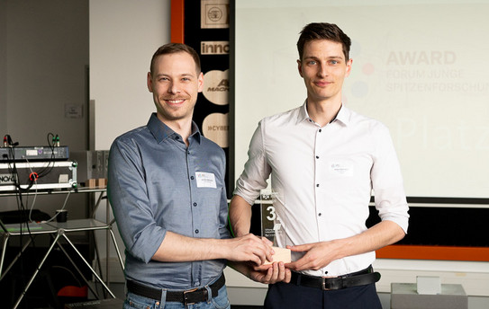 Foto: Zwei Männer in Hemden lächeln in die Kamera und halten eine Trophäe in der Hand, auf der die Zahl 3 zu sehen ist.