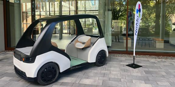 NeMo.bil_Cab Ein offenes Elektro-Gefährt mit zwei gegenüberliegenden Sitzen steht ausgestellt vor einer gläsernen Gebäudefassade. Daneben steht ein Aussteller mit der Aufschrift "NEMO Paderborn".
