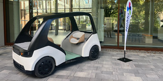 NeMo.bil_Cab An open electric vehicle with two seats facing each other is on display in front of a glass building facade. Next to it is an exhibitor with the inscription "NEMO Paderborn".