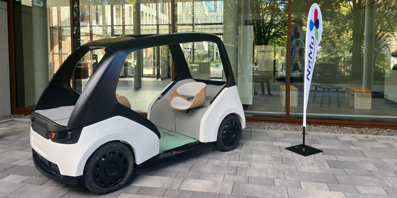 An open electric vehicle with two seats facing each other is on display in front of a glass building facade. Next to it is an exhibitor with the inscription "NEMO Paderborn".