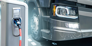 Elektrischer Schwerlastverkehr Ein grauer LKW steht neben einer elektrischen Ladesäule, in die ein elektrisches Autoladekabel eingesteckt ist.