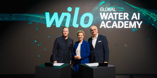 Auf dem Foto sieht man Oliver Hermes, Ina Brandes und Rektor Prof. Manfred Bayer vor einer digitalen Leinwand stehen. Sie stehen nebeneinander und lächeln für das Foto in die Kamera.