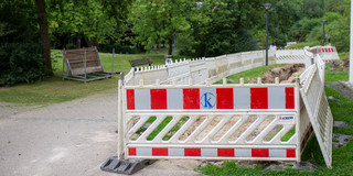 Neben einem Weg auf dem Campus ist ein längeres Stück Erde ausgehoben und mit rot-weißen Baustellenbegrenzungen eingezäunt. Im Hintergrund eine grüne Wiese mit Bäumen.