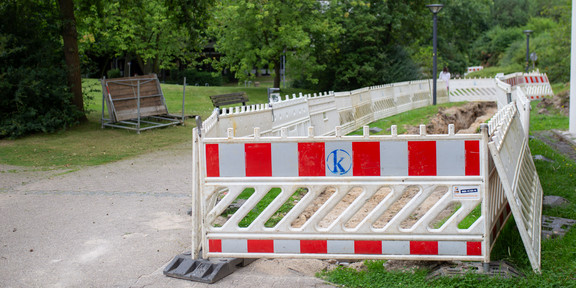 Next to a path on the campus, a stretch of earth has been excavated and fenced off with red and white construction site boundaries. In the background is a green meadow with trees.