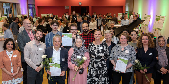 Ein Gruppenfoto von den verschiedenen Menschen mit Prof. Tessa Flatten vor einem gefüllten Saal. Drei Personen halten Urkunden in den Händen. Ein Mann mit grauen Haaren und Brille hält neben der Urkunde auf eine weiße Rose. Eine Frau in grau-schwarzem Hijab mit geometrischem Muster und eine Frau mit Brille und kurzen braunen Haaren halten jeweils eine weiße Rose.