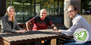 Two men and a woman sit outside at a table on the campus of the TU Dortmund.