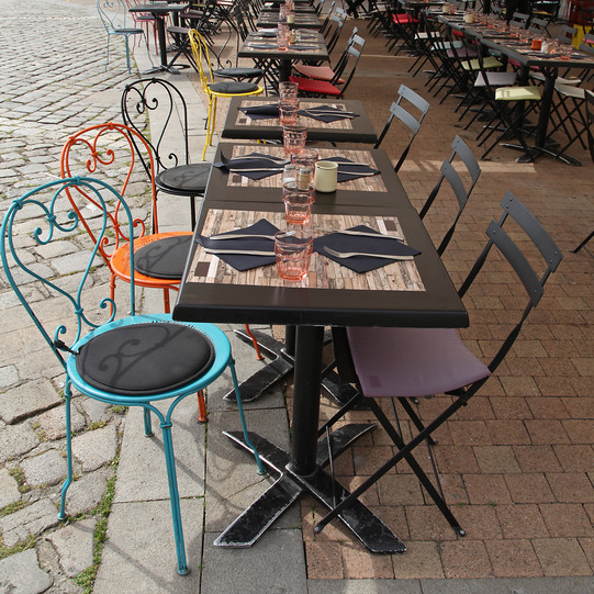 On a pavement there are three small tables with colourful chairs around them. 