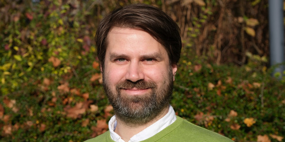 Zu sehen ist ein Porträtfoto von Nils Krüger vor einem waldig herbstlichen Hintergrund. Er trägt einen hellgrünen Pullover mit weißem Kragen.