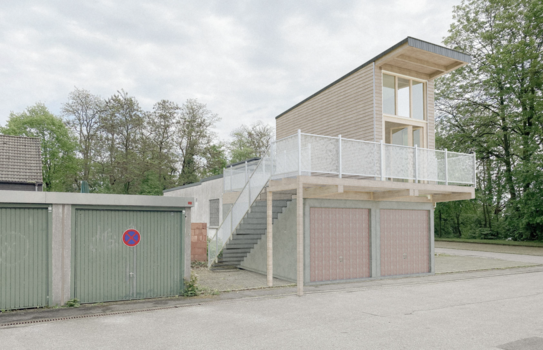 Mehrere Garagen auf denen ein modernes Tiny House gebaut ist. Das Tiny House hat große Fenster und eine Veranda mit weißem Geländer. Eine Treppe führt an der Seite der Garage zum Tiny House.