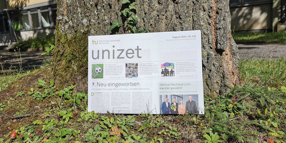 The picture shows the UNIZET edition leaning against a tree stump. Part of a building can be seen in the background.