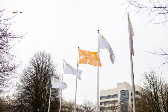 Fünf Fahnen an Fahnenmasten. Vier Fahnen sind weiß mit TU Dortmund Logo. Die mittlere Fahne ist orange. 