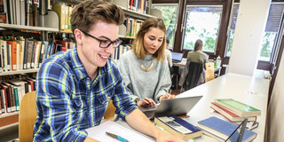 Zwei Studierende sitzen mit Lernmaterialien an einem Tisch in einer Bibliothek.