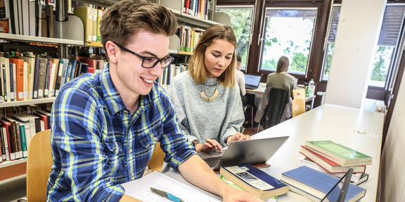 Lernen in der Bibliothek Zwei Studierende sitzen mit Lernmaterialien an einem Tisch in einer Bibliothek.