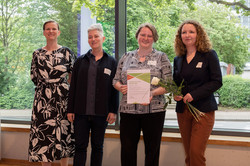 Ein Gruppenfoto von drei Frauen. Die erste von ihnen hat kurze graue Haare und trägt einen schwarzen Blazer. In der Mitte steht eine Frau mit kurzen braunen Haaren und Brille, die eine Urkunde vor sich hält. Die dritte Frau hat lockige dunkelblonde Haare und hält eine weiße Rose. Links von ihnen steht Prof. Tessa Flatten, eine blonde Frau im schwarzen Kleid mit abstrakten weißen Blüten.