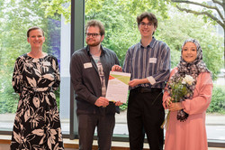Ein Gruppenfoto von zwei jungen Männern, die eine Urkunde zwischen sich halten, und einer jungen Frau mit grau-schwarzem Hijab mit geometrischem Muster, die eine weiße Rose hält. Links von ihnen steht Prof. Tessa Flatten, eine eine blonde Frau im schwarzen Kleid mit abstrakten weißen Blüten.