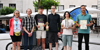 Foto: Eine Gruppe von sechs Menschen steht auf einem Platz in der Dortmunder Innenstadt. Sie halten Urkunde und Trophäen in den Händen.