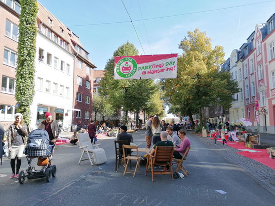 Mehrere Personen sitzen um einen Tisch herum auf einer Straße, rechts und links sind Häuser mit Bäumen und es ist Sommer.