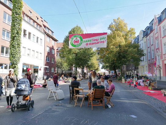 Several people are sitting around a table on the street. To the right and left are houses with trees. It is summer.