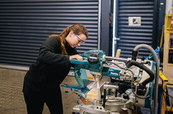Eine Frau in schwarzer Kleidung arbeitet an einer Kreissäge. Sie trägt eine Schutzbrille.
