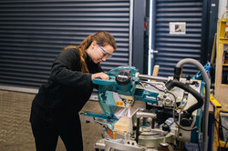 Eine Frau in schwarzer Kleidung arbeitet an einer Kreissäge. Sie trägt eine Schutzbrille.