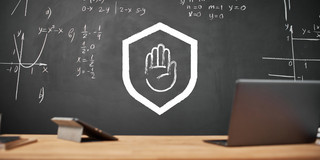 In the foreground are a laptop and a tablet on a table, in the background is a blackboard. On it are stylized form of an outstretched hand in a protective shield.
