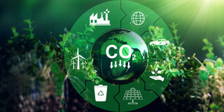 The photo shows a circular arrangement with the word CO2 in the center, from which arrows point downwards. The word is surrounded by various symbols such as a windmill, a car and a garbage can. In the background there are some plants and a shining sun at the top right of the image.