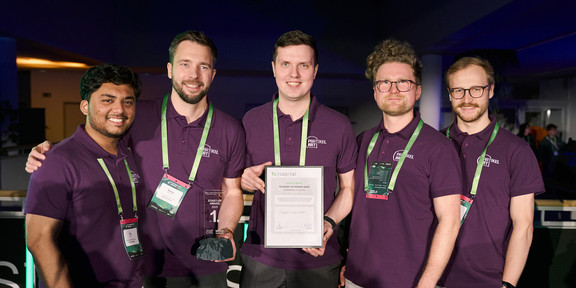 Foto: Fünf Personen in lila Poloshirts stehen zusammen, wobei eine Person eine Urkunde und eine andere eine Auszeichnung hält.