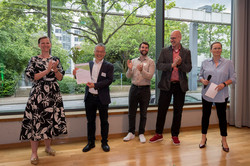 Ein Gruppenfoto, bei dem drei Männer in der Mitte stehen. Der erste Mann hat kurze graue Haare und eine Brille. Er hält eine Urkunde und eine weiße Rose neben sich. Der zweite Mann hat einen kurzen Bart und applaudiert. Der dritte Mann hat keine Haare und applaudiert ebenfalls. Links von ihnen steht Prof. Tessa Flatten, eine blonde Frau im schwarzen Kleid mit abstrakten weißen Blüten. Rechts von ihnen steht Silke Viol, eine blonde Frau in blauer Bluse mit Mikrofon in der Hand.