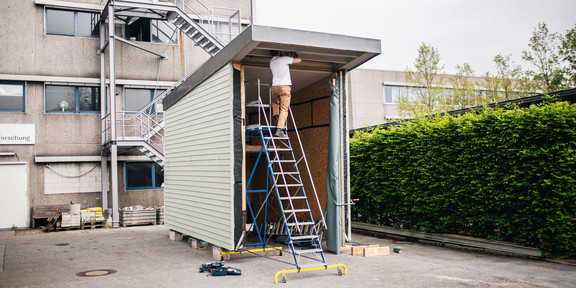 A person works on a ladder on an incomplete tiny house. The building has green walls and a flat roof. It stands on a concrete floor. 