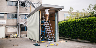 Eine Person arbeitet auf einer Leiter an einem unvollständigen Tiny-House. Das Gebäude hat grüne Wände und ein flaches Dach. Es steht auf Betonboden. 