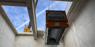 Photo: A large technical component is being lifted by a crane through an opening in a concrete building.