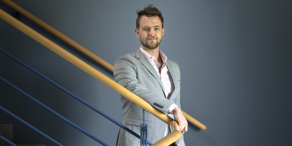 Eine Portraitaufnahme eines Mannes auf einer Treppe mit einem geschwungenen Geländer aus hellem Holz.
